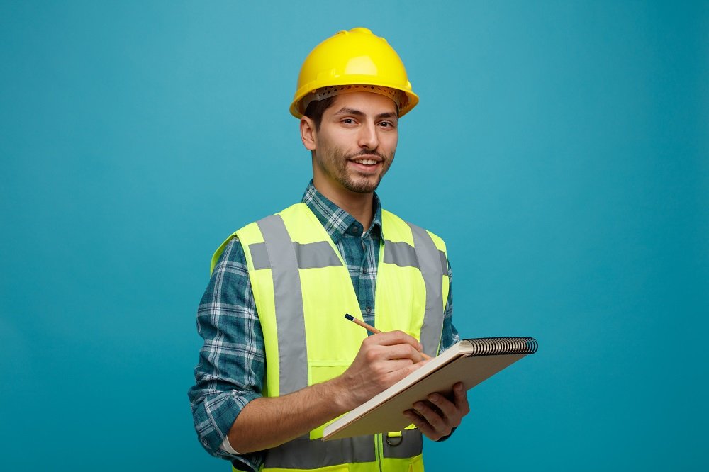 smiling young male engineer wearing safety helmet and uniform holding pencil and note pad looking at camera isolated on blue background